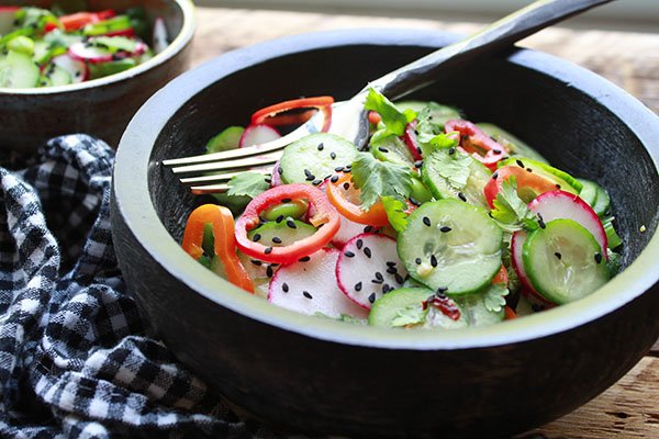 A black bowl filled with an Asian cucumber salad with lots of crunchy vegetables and sprinkled with black sesame seeds and topped with a fork with a black and white checkered napkin on the side.