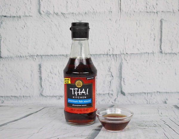 A bottle of fish sauce and a small glass bowl filled with fish sauce placed on top of a white plank board with white brick in the background.