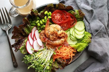 Grilled miso shrimp on top of greens with cucumbers, tomatoes, edamame pods, radishes and carrots with a grey napkin and silver fork on the side and miso dressing in a clear glass in the background.