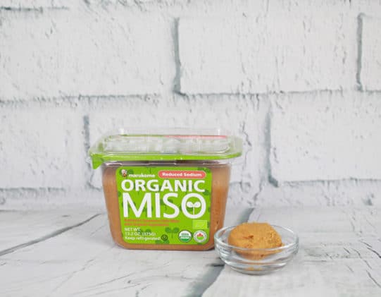 a container of organic miso paste on a white board with a tablespoon of paste in a glass bowl on the side