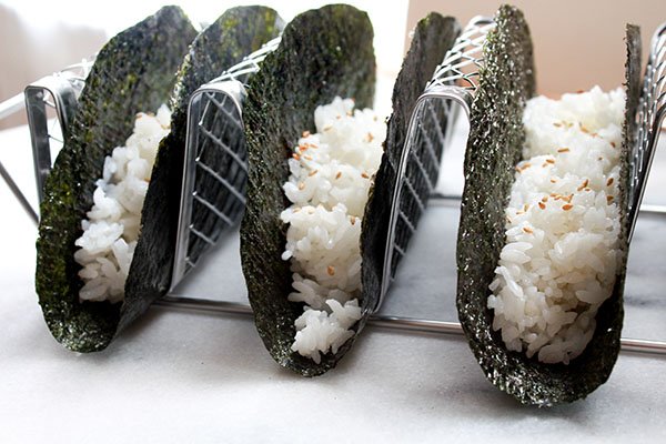 Three nori shells in a silver taco holder with white  rice nestled inside each shell.