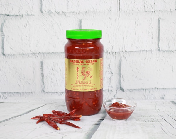 A bottle of sambal oelek and a small glass bowl filled with sambal oelek placed on top of a white plank board with white brick in the background.
