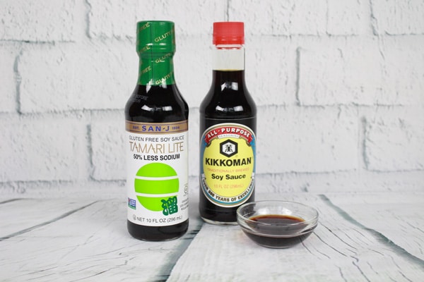 Two bottles of soy sauce and a small glass bowl filled with soy sauce placed on top of a white plank board with white brick in the background.