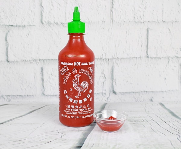 A bottle of sriracha chili sauce and a small glass bowl filled with sriracha placed on top of a white plank board with white brick in the background.