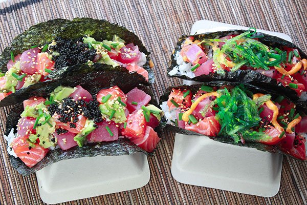 Beautiful sushi-grade tuna and salmon cubes nestled inside a nori shell on top of white rice and topped with black caviar and wakame seaweed salad placed in taco holders on top of a bamboo placemat.