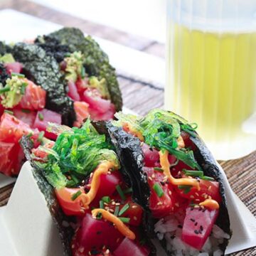 Beautiful sushi-grade tuna and salmon cubes nestled inside a nori shell on top of white rice and topped with black caviar and wakame seaweed salad placed in taco holders on top of a bamboo placemat with a frosty mug of beer in the background.