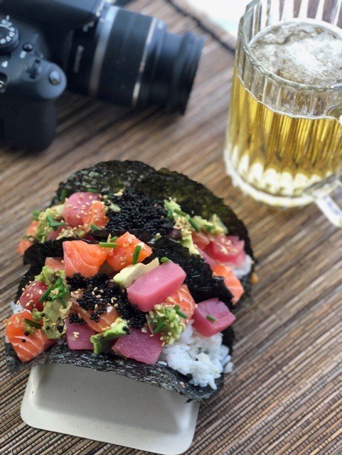 A sushi taco on a bamboo mat with a frosty beer and a professional camera in the background.