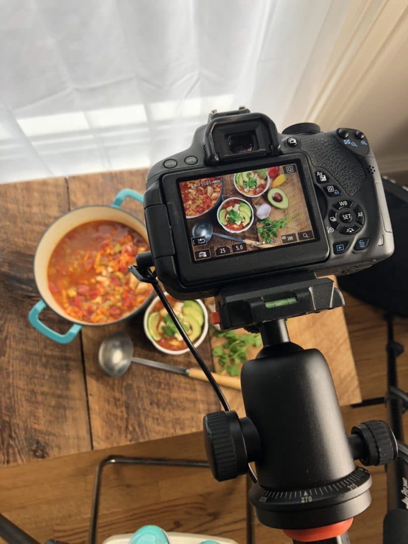 A professional camera shooting down on a large bowl of soup with a ladel on top of a wooden board.