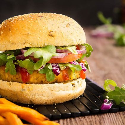 A thick vegetarian tofu burger sandwiched in between a burger bun and topped with greens, tomatoes, and red onions on top of a black plank board with fries on the side.