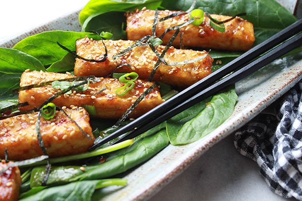 Sesame seared tofu strips on a bed of spinach topped with sesame seeds and seaweed strips presented on a long gray serving plate with black chopsticks on the side.