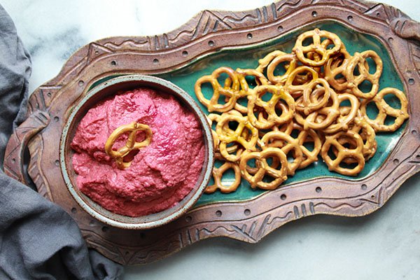red beet hummus in a bowl with a side of pretzels for dipping on top of a green serving plate