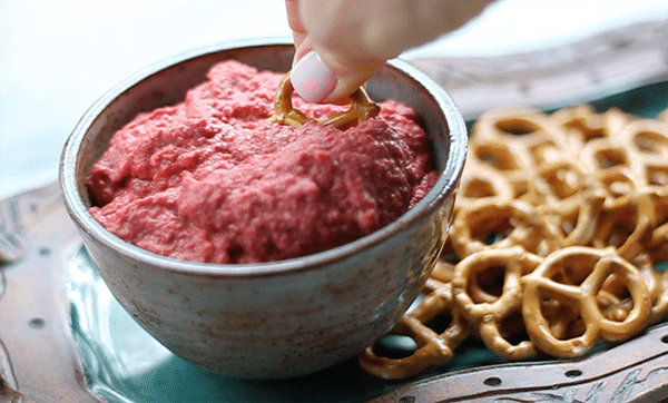red beet hummus with a pretzel being dipped into a bowl with a side of pretzels on top of a green serving plate