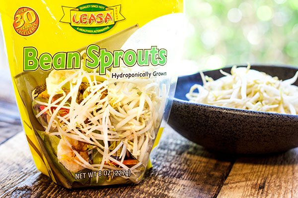 A package of bean sprouts on a wooden board with a black bowl of bean sprouts on the side.