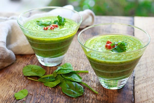 two clear bowls of chilled green gazpacho soup on top of a wooden board with fresh spinach on the side.