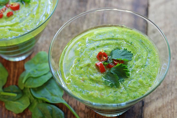 green gazpacho soup in two clear class bowls with fresh spinach on this side on top of a wooden board