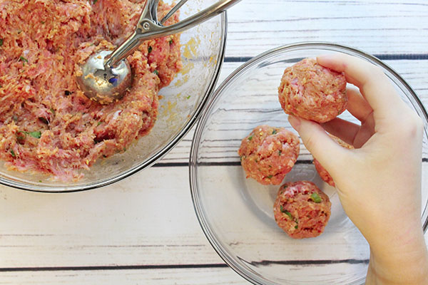 A woman's hand forming meatballs above a glass mixing bowl with already formed meatballs inside the bowl, and a large glass mixing bowl on the side with the meatball mixture.