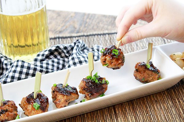 A woman's hand picking up a skewered sweet and spicy BBQ meatball above a row of meatballs placed on a thin white rectangular serving platter with a mug of beer and a checkered napkin on the side.