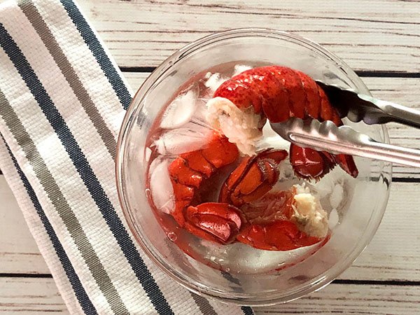 Three cooked Maine lobster tails sitting in an ice bath in a clear glass bowl with tongs holding one lobster tail on top of a white plank board with a striped napkin on the side.