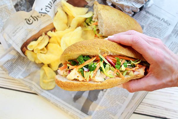 A woman's hand holding a lobster banh mi sandwich above a board with potato chips on top of a newspaper.