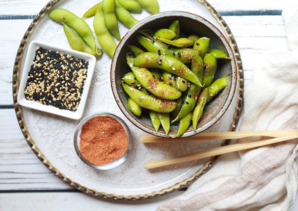 A small bowl of spicy edamame on a round white plate with a bowl of sesame seeds, sriracha seasoning, and wooden chopsticks on the side. 
