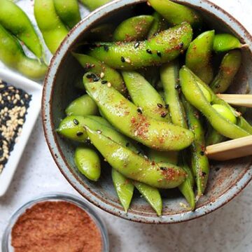 A small bowl of spicy edamame pods with chopsticks, and sesame seeds and sriracha powder on the side.