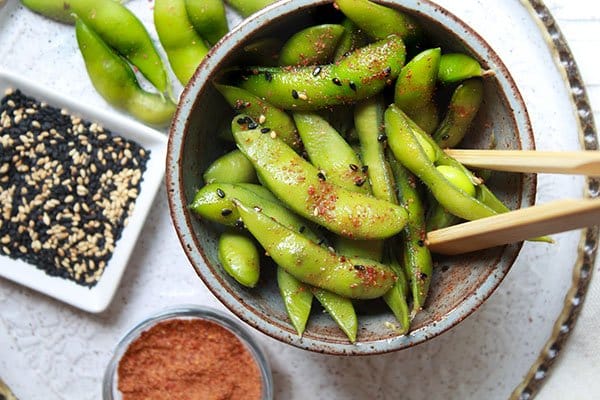 A small bowl of spicy edamame pods with chopsticks, and sesame seeds and sriracha powder on the side.