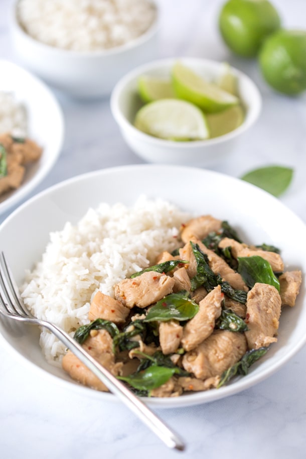 Thai basil chicken in a white bowl with white rice and fresh lime wedges in the background.