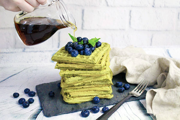 A  woman's hand pouring maple syrup on top of a stack of matcha green tea waffles topped with fresh blueberries placed on top of a black slate tray with a fork and linen napkin on the side.