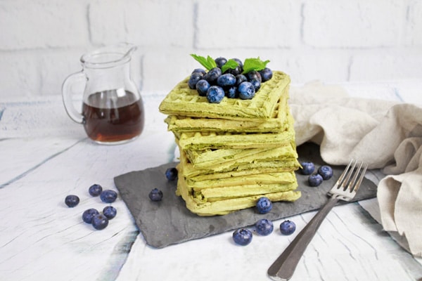 A stack of matcha green tea waffles topped with fresh blueberries placed on top of a black slate tray with a glass pourer filled with maple syrup on the side.