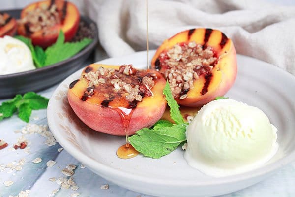 grilled peaches on a white plate topped with a five spice oats crumble and a scoop of vanilla ice cream on the side