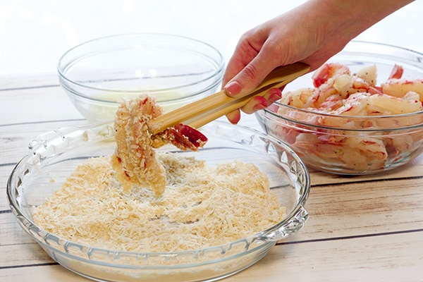 A woman's hand dipping a butterflied shrimp into a Panko bread crumb mixture, with a glass bowl of raw shrimp on the side.