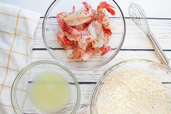 Three glass prep bowls filled with ingredients for baked coconut shrimp on top of a white board with a whisk and napkin on the side.