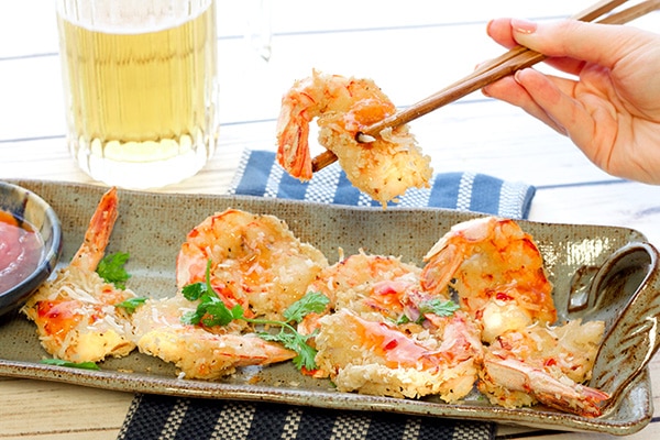 A woman's hand holding chopsticks with a baked coconut shrimp over a platter of coconut shrimp with a beer in a mug on the side.