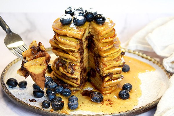 A high stack of pumpkin chocolate chip pancakes topped with fresh blueberries on top of a white plate with a fork and napkin on the side placed on top of a marble surface.