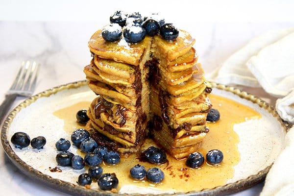 Fluffy pumpkin pancakes stacked high on a plate dripping with maple syrup and topped with fresh blueberries and powdered sugar.