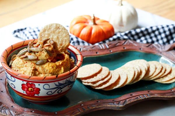 A small dipping bowl filled with pumpkin hummus topped with pumpkin seeds served on top of a green platter with rice crackers on the side and a black and white checkered napkin on the side.