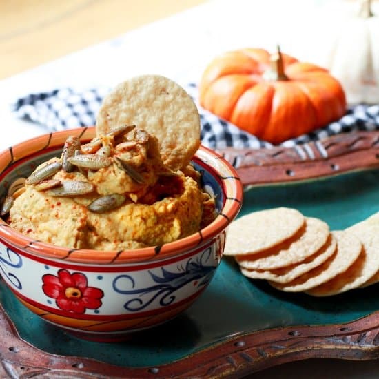 Pumpkin hummus in a decorative bowl with rice crackers on the side a baby pumpkin in the background.