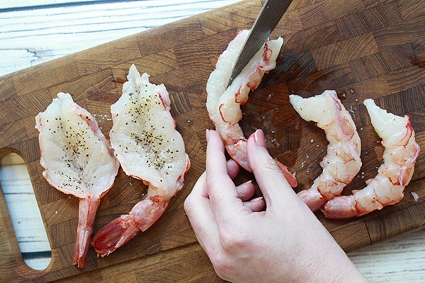 a woman holding a knife while butterflying several jumbo shrimp on a wooden cutting board.