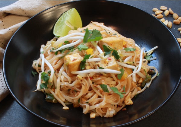 tofu pad thai in a black bowl garnished with a lime wedge with a napkin on the side