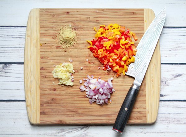 Chopped multi-colored bell peppers, grated ginger, sliced garlic, and chopped red onions on top of a wooden cutting board with a chef's knife placed on the side on top of a white wooden plank board.