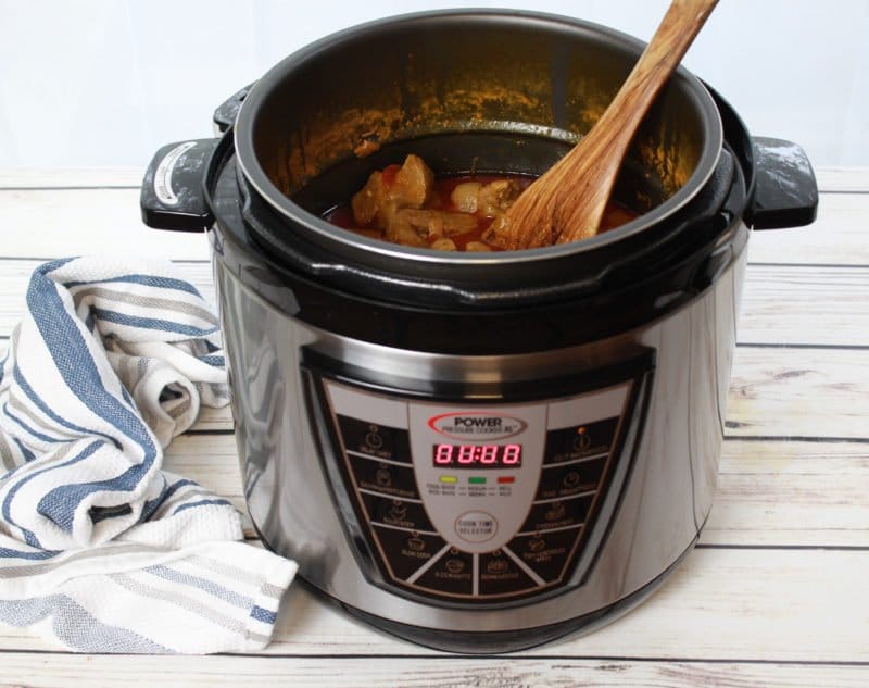 An Instant Pot with dinner being prepared placed on top of a white wooden board with a kitchen napkin on the side.