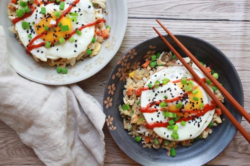 A gray bowl filled with cauliflower fried rice with crab topped with a fried egg and a pair of chopsticks on top of a wooden board with another bowl of fried rice behind it.