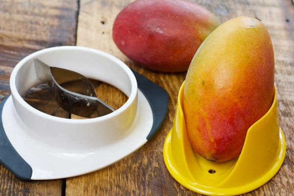 Whole fresh mangos on top of a wooden board with a mango slicer on the side.