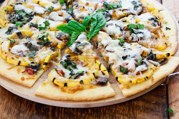 Thin-Crust Delicata Squash and Prosciutto Pizza topped with sweet roasted squash, shallots, mushrooms, and melty cheese on top of a pizza stone with fresh basil leaves on the side placed on top of a wooden board.