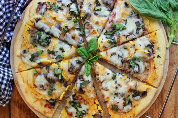 Thin-Crust Delicata Squash and Prosciutto Pizza topped with sweet roasted squash, shallots, mushrooms, and melty cheese on top of a pizza stone with fresh basil leaves on the side placed on top of a wooden board.