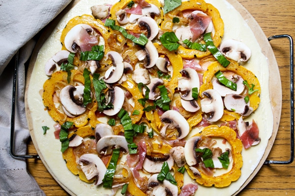 A delicata squash pizza with lots of toppings on top of a pizza stone ready to be placed in the oven for baking.