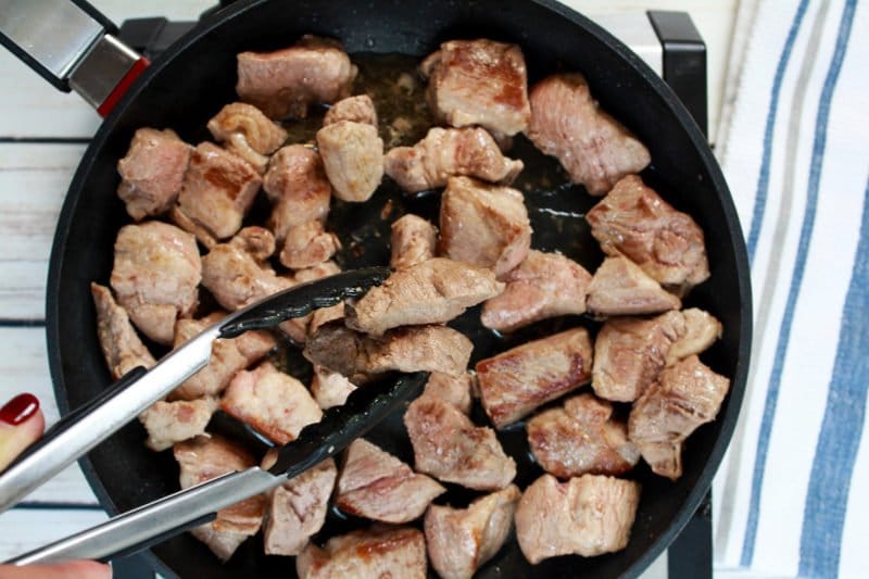 Chunks of pork butt pieces being seared in a large frying pan with a kitchen towel on the side.
