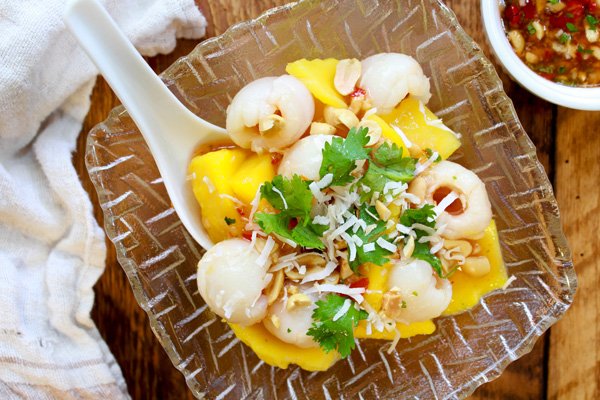 A tropical mango and lychee salad in a glass serving bowl with a large white spoon inserted on top of a wooden board with a small white bowl of spicy sauce on the side.