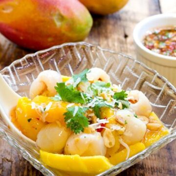 A tropical mango and lychee salad in a glass serving bowl with a large white spoon inserted on top of a wooden board with a small white bowl of spicy sauce on the side and two whole mangos place behind.