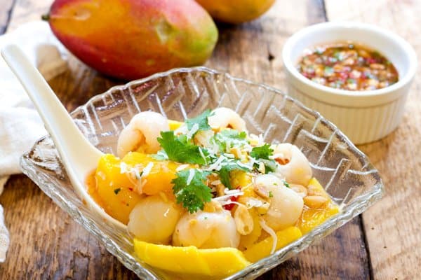 A tropical mango and lychee salad in a glass serving bowl with a large white spoon inserted on top of a wooden board with a small white bowl of spicy sauce on the side and two whole mangos place behind.